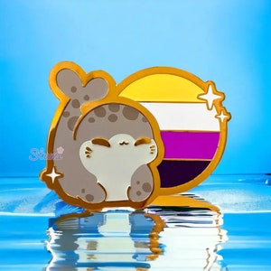 May include: Gold-tone enamel pin featuring a cartoon seal with a closed-eye smile, resting against a circular background with the colors of the non-binary pride flag. The flag is yellow, white, purple, and black. The pin has two small gold stars on the right side.