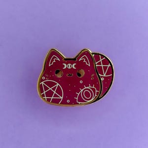 May include: A burgundy cat-shaped enamel pin with gold trim. The pin features white occult symbols, including a pentagram, eye, and crescent moons. The pin is set against a purple background.