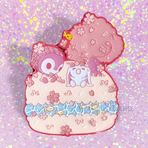 May include: A pink and white enamel pin featuring two cartoon characters, a bunny and a penguin, under a pink cherry blossom tree. The characters are sitting on a pink and white cake with blue water and yellow fish.