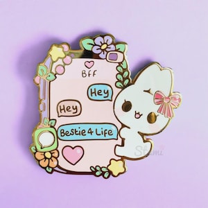 May include: A gold-tone enamel pin featuring a cartoon bunny holding a pink and white phone with the text "Bff", "Hey", and "Bestie 4 Life" on the screen. The phone is surrounded by flowers and stars.