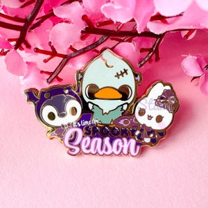 May include: A gold enamel pin with three cartoon characters: a purple bat, a Frankenstein-themed duck, and a white bunny. The pin has the text "It's time for Spooky Season" in purple and white. The background is pink.