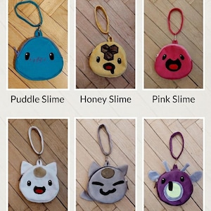 Puede incluir: Colección de seis monederos de Slime Rancher. Cada monedero tiene un diseño y color diferentes, con personajes como Puddle Slime, Honey Slime y Pink Slime. Cada monedero tiene una cremallera y una correa de muñeca. El texto "Slime Rancher Coin Purse Collection" está en la parte superior.