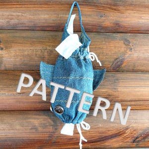 May include: A blue denim shark-shaped bag with a white drawstring and a button on the front. The bag is hanging from a wooden surface.