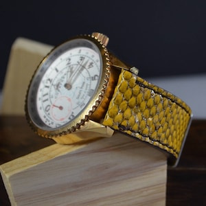 Pode incluir: Um relógio de pulso dourado com um mostrador branco e marcas pretas. O relógio tem uma pulseira de couro amarela com um padrão texturizado.