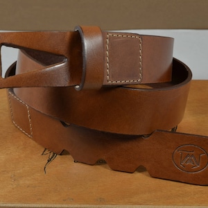 May include: A brown leather belt with a buckle featuring a unique design. The belt is folded in half and the buckle is visible. The belt is on a wooden surface.