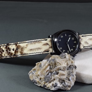 May include: A black watch with Roman numeral markings on the face and a white leather band with a snakeskin pattern.