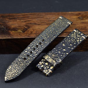 May include: Two-piece watch strap set. The straps are black with a gold, textured pattern. The straps are made of a material that resembles stingray leather. The straps are displayed on a dark wooden block.