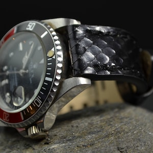 Pode incluir: Um relógio de pulso preto e vermelho com uma pulseira de couro preto. O mostrador do relógio tem marcas brancas e um fundo preto. A pulseira é feita de um material texturizado.