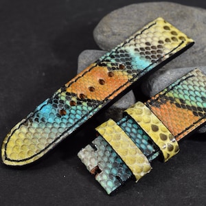 Könnte beinhalten: Ein zweiteiliges Uhrenarmband mit einem mehrfarbigen Schlangenledermuster in Gelb-, Orange- und Türkistönen. Das Armband hat schwarze Nähte an den Kanten, ein spitz zulaufendes Ende am längeren Teil und zwei verschiebbare Schlaufen am kürzeren Teil.
