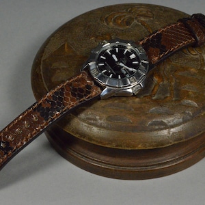 Op de afbeelding: Een zilveren polshorloge met een zwarte wijzerplaat en een bruin leren bandje met een slangenhuidpatroon. Het horloge ligt op een houten doos met een gesneden ontwerp.