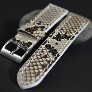 May include: A white leather watch strap with a brown snakeskin pattern. The strap has a silver buckle and is stitched with white thread.