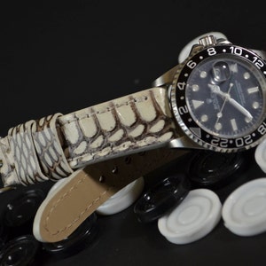 Può includere: Un cinturino da orologio in pelle bianca con un motivo a coccodrilo. Il cinturino è attaccato a un orologio argentato con un quadrante nero e una lunetta nera con numeri bianchi.