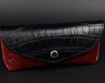 May include: A black leather glasses case with a crocodile pattern and a red leather lining. The case has a single button closure.