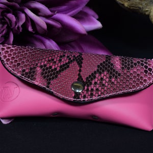 Könnte beinhalten: Ein rosafarbenes Leder-Brillenetui mit Schlangenhautmuster auf der Klappe. Das Etui hat einen silbernen Druckknopfverschluss und ein kleines Logo auf der Vorderseite.
