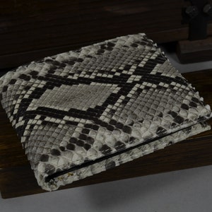 Pode incluir: Uma carteira de pele de cobra cinza e preta com um padr&atilde;o texturizado. A carteira est&aacute; dobrada e est&aacute; sobre uma superf&iacute;cie de madeira escura.