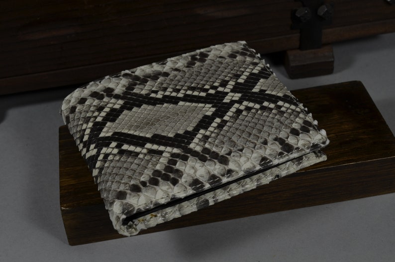 Pode incluir: Uma carteira de pele de cobra cinza e preta com um padr&atilde;o texturizado. A carteira est&aacute; dobrada e est&aacute; sobre uma superf&iacute;cie de madeira escura.