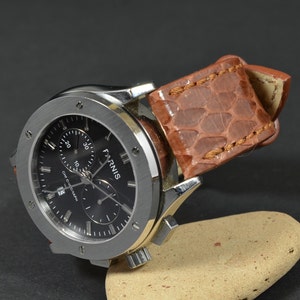 Puede incluir: Un reloj de pulsera cronógrafo plateado con una esfera negra y una correa de cuero marrón. La esfera del reloj tiene marcas blancas y la palabra "FARNIS" impresa en ella.