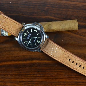 May include: A brown leather watch strap with a textured pattern. The strap is attached to a silver watch with a black face and white markings. The watch face reads "Luminor Marina Panerai".