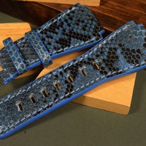 Puede incluir: Una correa de reloj de piel de serpiente azul y negra con detalles de costura azul.