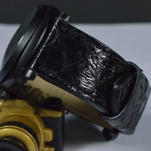 Peut inclure: Une montre-bracelet noire avec un bracelet en cuir texturé. Le cadran de la montre est sombre et le bracelet présente un motif. La montre repose sur un objet doré et noir.