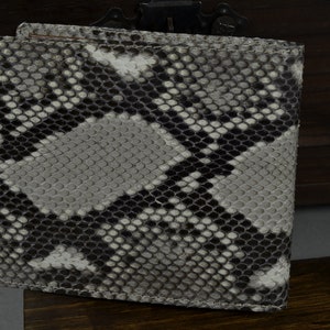 May include: A bi-fold wallet with a snakeskin pattern in shades of grey, black, and white. The wallet is made of a textured material and has a rectangular shape. The wallet is closed and appears to be new.
