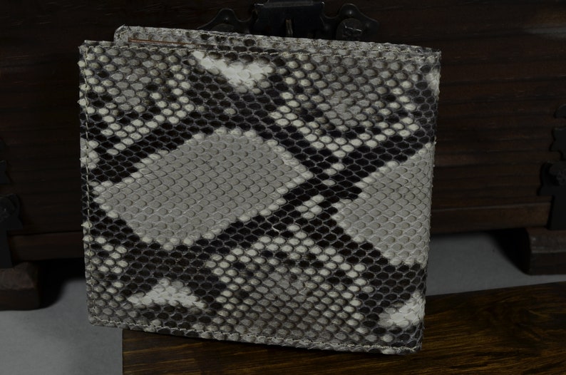 May include: A bi-fold wallet with a snakeskin pattern in shades of grey, black, and white. The wallet is made of a textured material and has a rectangular shape. The wallet is closed and appears to be new.