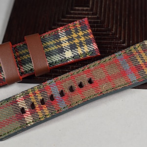May include: A red, green and brown tartan watch strap with brown leather keepers. The strap has dark green stitching around the edges.