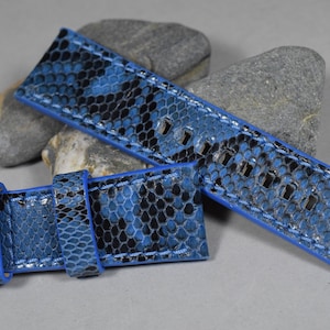 Peut inclure: Un bracelet de montre en cuir de serpent bleu et noir avec des détails de couture bleus. Le bracelet est présenté plié en deux.