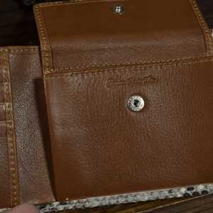 May include: Open brown leather wallet with a snakeskin exterior. The wallet is open, showing card slots and a snap closure. The interior is brown leather with a stamped logo.