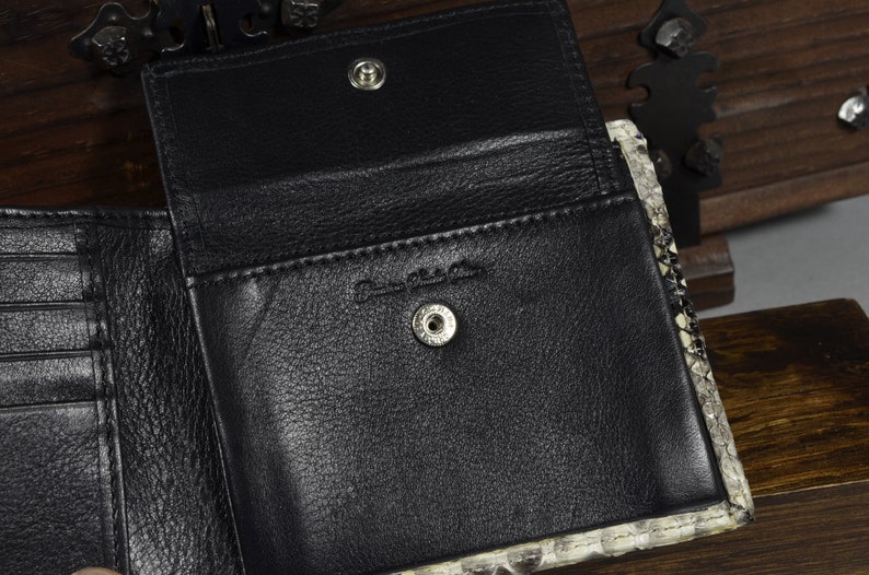 May include: A black leather wallet with a snakeskin pattern on the exterior. The wallet is open to reveal a black leather interior with multiple card slots and a snap closure.