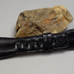 Op de afbeelding: Een zwarte horlogeband met een textuur die lijkt op die van een reptiel. De band heeft meerdere gaten voor verstelling en een gespsluiting. De achtergrond toont een stuk drijfhout.