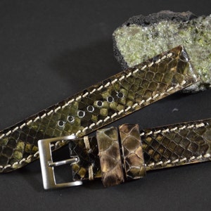 Può includere: Un cinturino da orologio in pelle di serpente marrone e verde con cuciture bianche. Il cinturino ha una fibbia argentata.