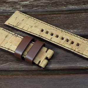 May include: A brown leather watch strap with a cork top and a stitched edge. The strap is shown on a wooden surface.