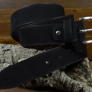 Peut inclure: Une ceinture en cuir noir avec une boucle argentée. La ceinture est pliée et présentée sur un morceau de liège.