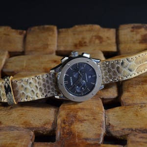 Puede incluir: Un reloj de pulsera cronógrafo plateado con esfera negra y correa de piel de serpiente marrón. El reloj está sobre una superficie de madera.