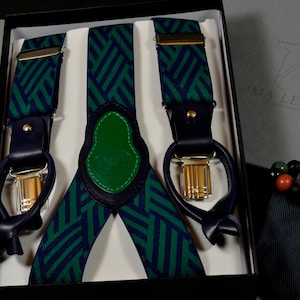 Può includere: Un paio di bretelle con motivo verde e blu con clip in metallo dorato e dettagli in pelle. Le bretelle sono in una scatola nera con interno bianco.