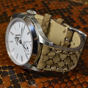 Peut inclure: Une montre-bracelet argentée avec un cadran blanc et des index en chiffres romains. La montre a un bracelet en cuir brun avec un motif texturé.