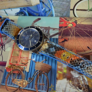 Pode incluir: Uma pulseira de relógio de couro com padrão azul e laranja com um detalhe de costura azul. A pulseira do relógio está enrolada em torno de um relógio azul e prateado com um mostrador azul.