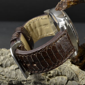 Könnte beinhalten: Ein braunes Lederarmband mit einem strukturierten, schlangenartigen Muster. Das Armband ist an einem silbernen Uhrengehäuse befestigt.