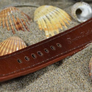 May include: A brown leather watch strap with stitching and holes for adjusting the size. The strap is lying on a bed of sand with seashells.