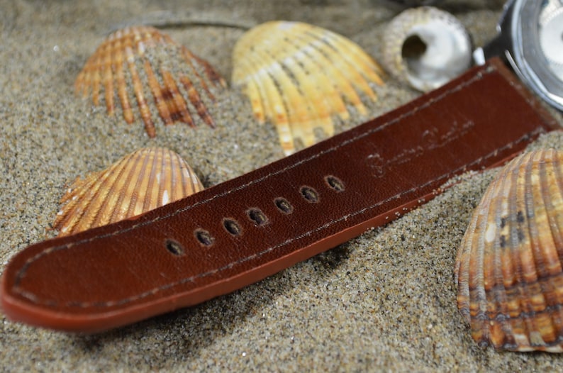May include: A brown leather watch strap with stitching and holes for adjusting the size. The strap is lying on a bed of sand with seashells.