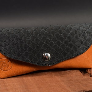May include: A brown leather glasses case with a black snakeskin pattern on the top. The case has a silver button closure and a logo on the front.