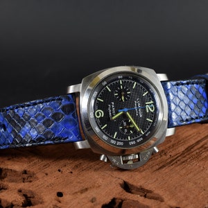 Pode incluir: Um relógio de pulso cronógrafo preto com uma pulseira de pele de cobra azul e preta. O mostrador do relógio tem um fundo preto com marcas brancas e uma pequena janela de data. O mostrador do relógio também tem o texto "Luminor Regatta" e "Panerai Flyback" impresso nele.