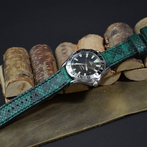 Op de afbeelding: Een groen leren horlogebandje met een gestructureerd slangenhuidpatroon. Het bandje is bevestigd aan een zilveren horloge met een zwarte wijzerplaat en een datumscherm.