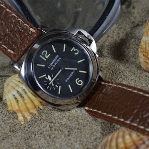 May include: A brown leather watch strap with white stitching, attached to a silver watch with a black face. The watch face has white markings and the text "Luminor Marina" and "Panerai". The watch is lying on a sandy surface with seashells.