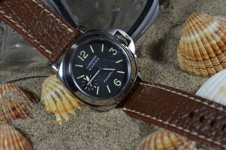 May include: A brown leather watch strap with white stitching, attached to a silver watch with a black face. The watch face has white markings and the text "Luminor Marina" and "Panerai". The watch is lying on a sandy surface with seashells.
