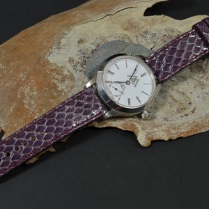 Puede incluir: Un reloj plateado con una esfera blanca y marcadores de hora romanos negros. El reloj tiene una correa de cuero morado con un patrón texturizado.