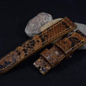 May include: Two-piece watch strap in a snakeskin pattern. The strap is brown with black and tan accents. The watch band is made of a textured material and has a light brown underside. The strap is shown on a black surface.