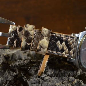 Pode incluir: Uma pulseira de relógio de pele de cobra castanho e bege com uma fivela prateada. A pulseira é feita de couro genuíno e tem um padrão texturizado único.