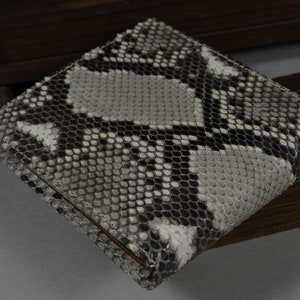 May include: A bi-fold wallet crafted from genuine snakeskin. The wallet displays a pattern of grey, black, and white scales. The rectangular wallet appears to be unused.